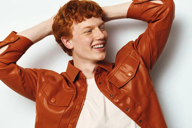 Young man with red hair wearing brown leather jacket and white shirt smiles happily against white background. Casual style, portrait with hands behind head, relaxed and joyful expression.