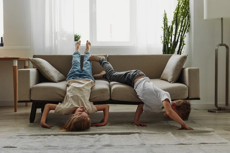 Two playful children in casual clothes balancing upside down on a sofa in a bright living room with natural light and green plants. Concept of fun, childhood, and active play.