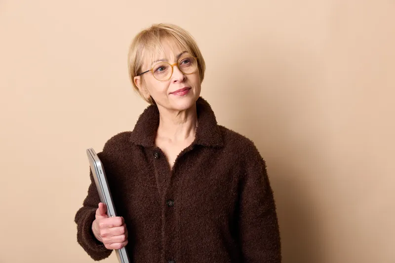 Senior woman with glasses holding a tablet, indoors portrait on beige background, calm smile, natural light, technology for communication and daily information