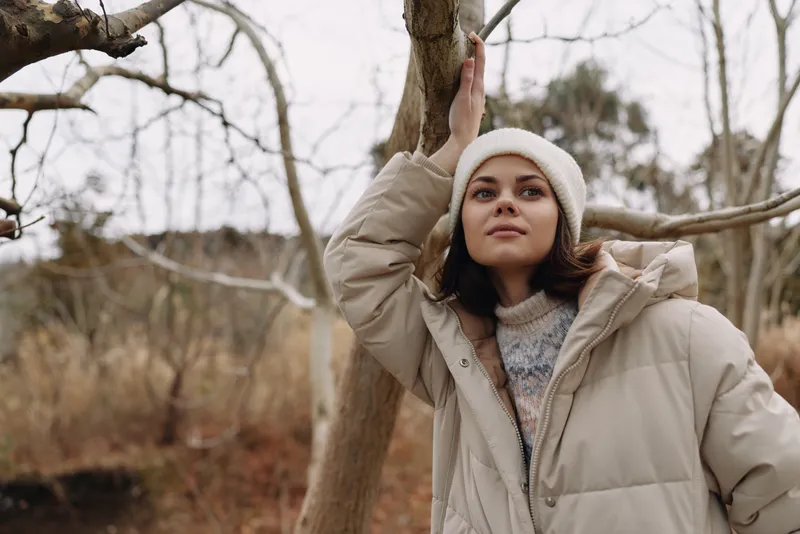 Woman in a beige puffer coat outdoors in winter, leaning against a tree branch, serene expression, soft natural light, cozy seasonal mood for lifestyle shooting, tranquil atmosphere