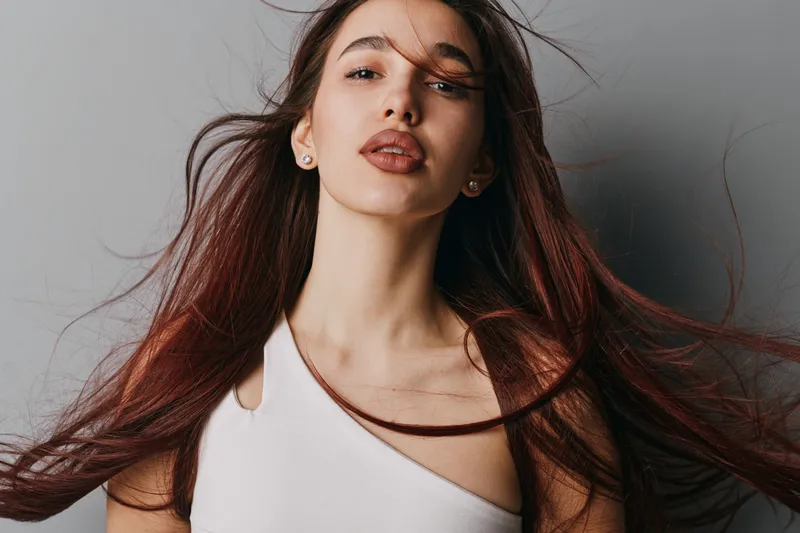 Young woman with long hair in white top posing against neutral background. Beauty portrait with natural makeup, confident expression and hair flowing for fashion or lifestyle concept.