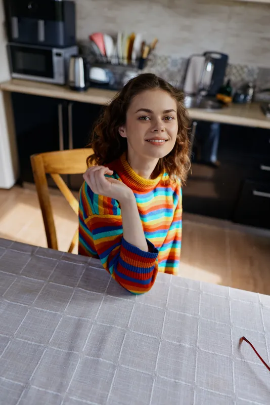 A woman sits at a kitchen table wearing a bright rainbow striped sweater, smiling softly, elbow resting on the table. A casual home setting conveys relaxed lifestyle and comfort in everyday moments.