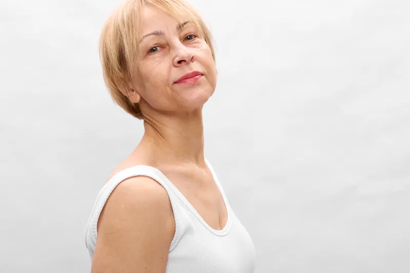Woman portrait elderly face profile studio calm white background, short blonde hair, relaxed expression, shoulders and neck visible, side view studio lighting, minimal monochrome look, copy space