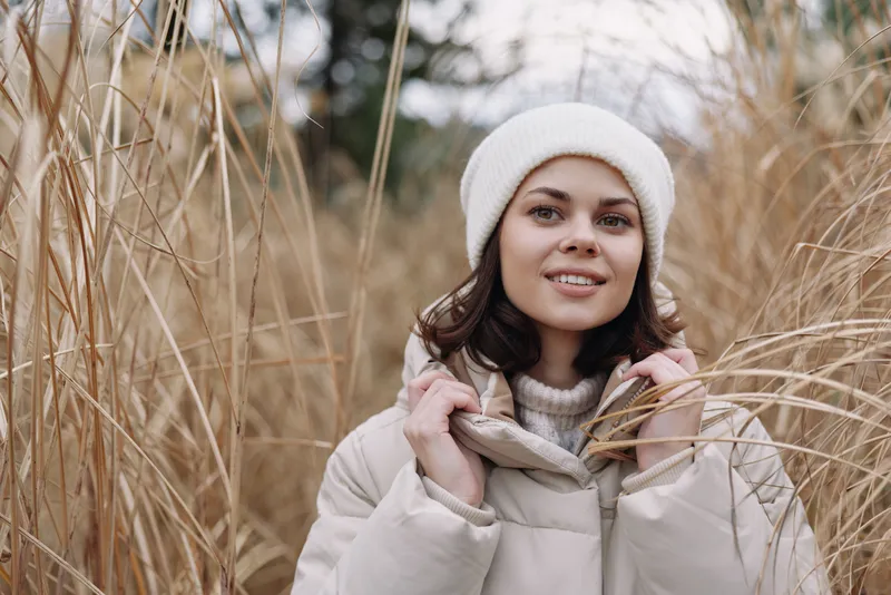 Winter outdoor portrait of a smiling woman in a beige coat and white beanie among tall reeds, capturing cozy, serene fashion and natural countryside mood.