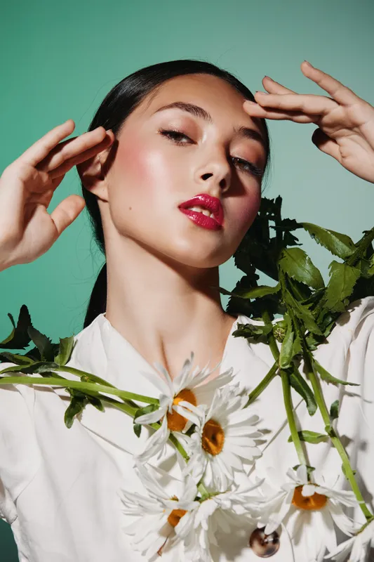 Close-up of a stylish young woman with flawless glossy skin and bold red lips environmental leaves and daisies draped over her shoulders on teal background. Fashion beauty concept.