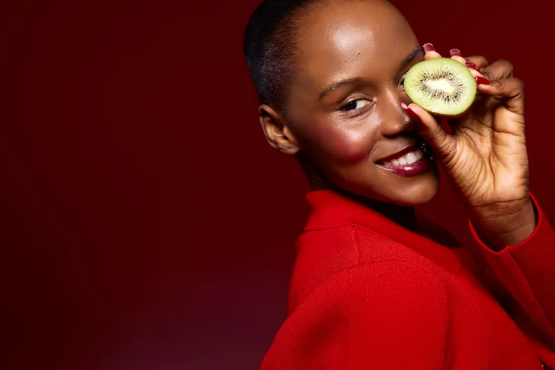 Portrait of a smiling African woman with dark skin wearing a red sweater holding a half kiwi fruit near her eye on red background. Concept of healthy lifestyle, natural beauty, and wellness.