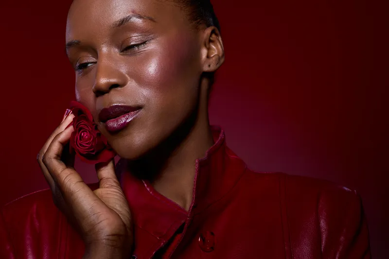 Close-up of a dark skinned African woman with closed eyes holding a red rose near her face. She wears a red leather jacket against a red background. Concept of beauty, elegance, and self-care.