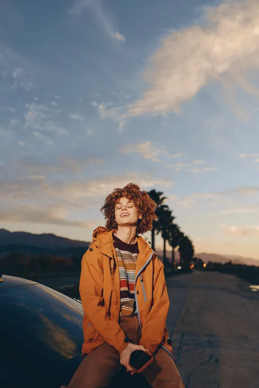Woman lifestyle outdoors at sunset, smiling with curly hair, wearing a jacket while sitting on a car hood, mountains and road in warm evening light, calm, slow smile