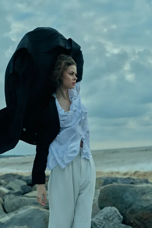 Woman on a windy beach holds a flowing coat aloft as stormy clouds loom, creating a dramatic outdoor fashion moment with rugged rocks and sea backdrop.