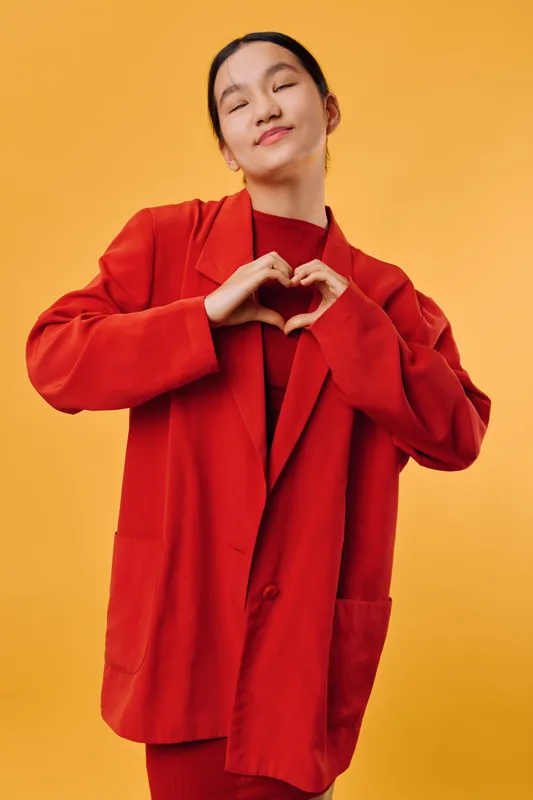 Young woman in red outfit makes heart shape with hands for love, affection and positive feelings on yellow background in studio. Female model shows gesture for kindness and care.