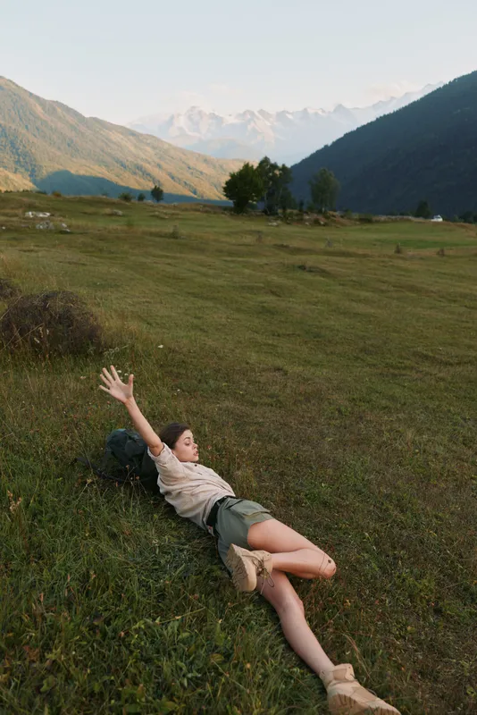 Relaxed woman lying on a grassy meadow with distant mountains, enjoying sunshine and fresh air, captured as a casual outdoor lifestyle moment in a natural landscape setting.