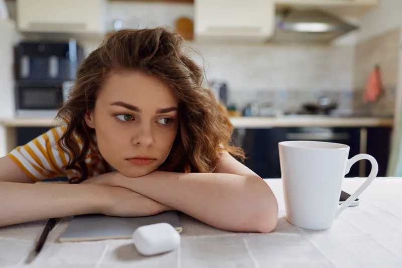 A young woman rests her head on folded arms at a sunlit kitchen table, a white mug and a remote nearby, conveying fatigue, burnout and introspection in a cozy home setting.
