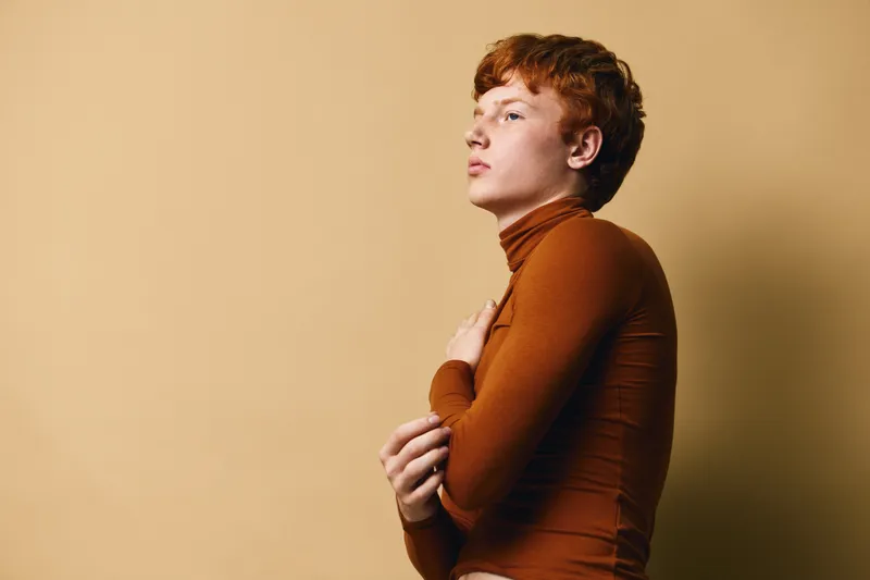Portrait of thoughtful redhead young man wearing brown turtleneck sweater against beige background with arms crossed, looking sideways in studio lighting.