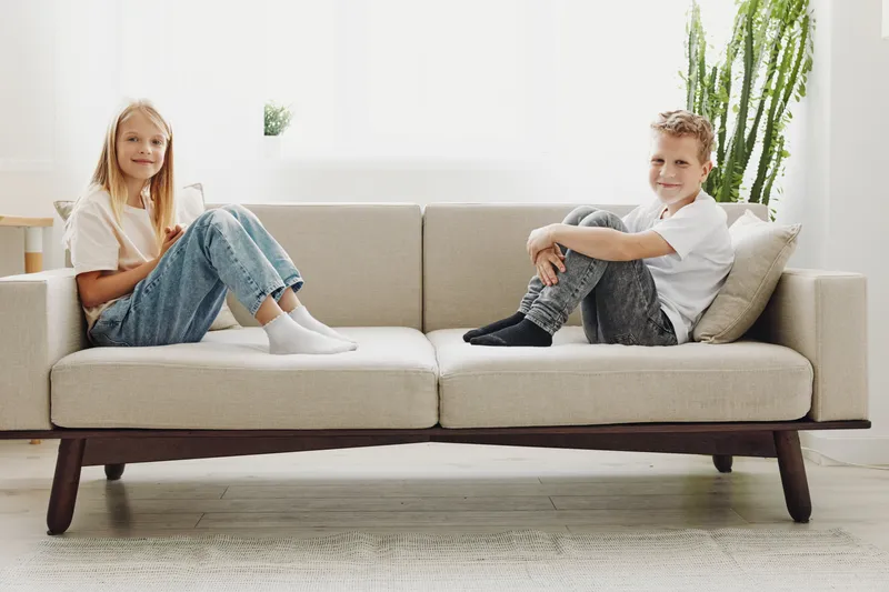 Two children sit on opposite ends of a modern beige sofa in a bright living room with a tall green plant and white walls. Concept of family, home, and minimalism.
