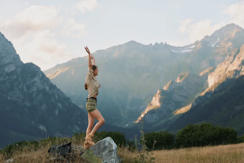 Woman outdoors on a rock with arms raised in a mountain valley. Active lifestyle, adventure, and vitality in nature. Bright natural light highlights confidence and freedom, and adds a sense of