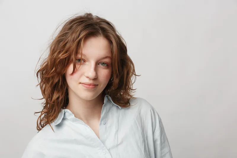 Young woman with curly hair and light blue shirt smiles gently against a plain white background. Natural beauty portrait with soft lighting and casual style concept.