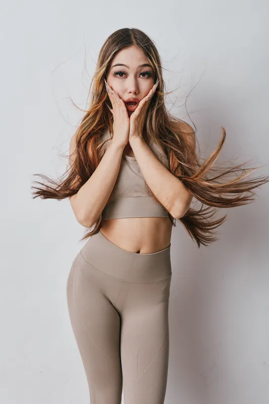 Woman in beige sportswear shows surprised emotion with hands on cheeks and flowing hair in studio for fitness, health and lifestyle concept on white background.