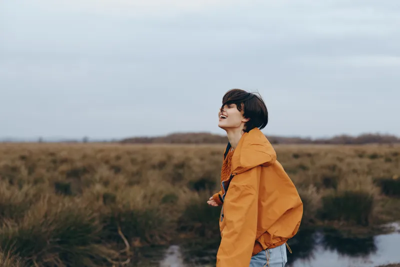 Woman lifestyle outdoors portrait, standing by river, wearing orange hooded jacket, short dark hair, relaxed smile, cloudy countryside, nature field, calm water, soft light