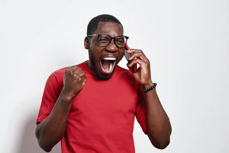 African American man with dark skin wearing glasses and red shirt talking on phone with excited expression and fist pump for success or good news on white background.