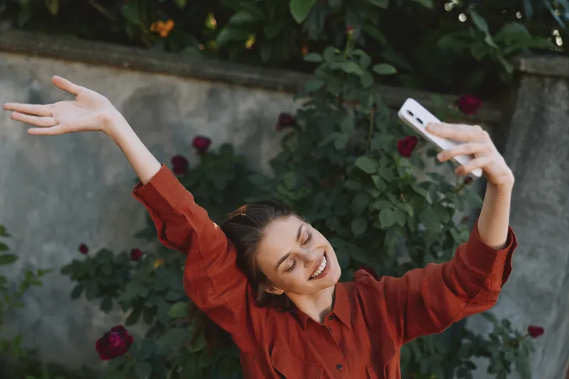 Young woman with brunette hair in a red blouse smiles and stretches her arms in a joyful selfie amid a garden filled with roses and greenery, bathed in soft natural light