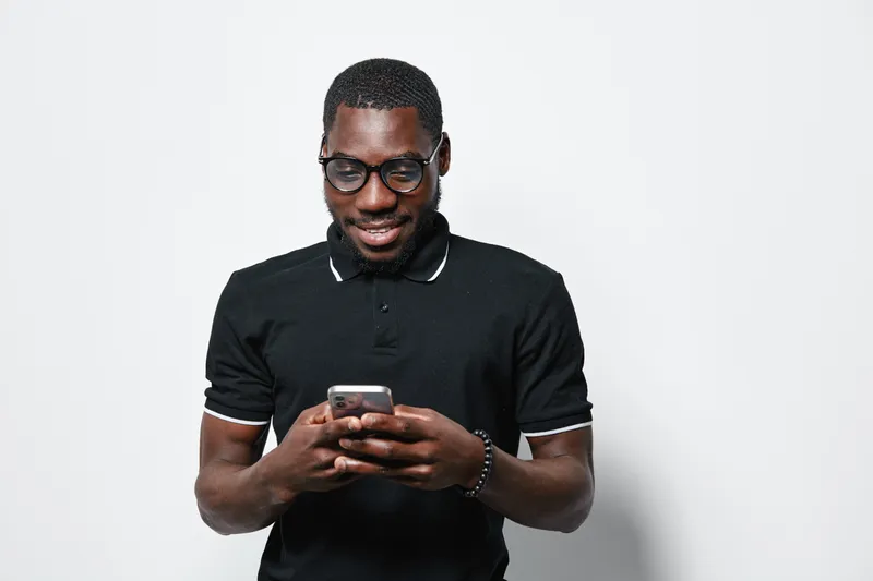 African American man with dark skin and glasses wearing black polo shirt using smartphone and smiling on white background. Casual, technology, communication and lifestyle concept.