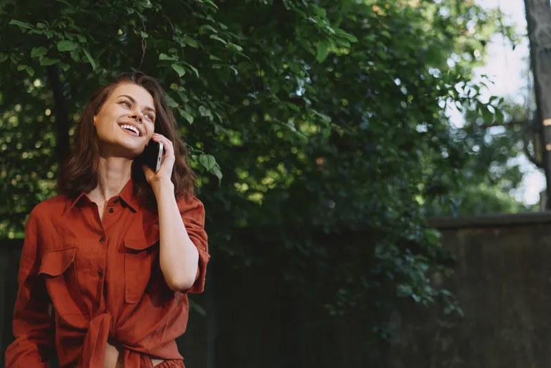 Young woman in a red shirt smiles softly while talking on a cellphone in a sunlit garden, surrounded by lush greenery and trees, radiating a warm casual outdoor mood