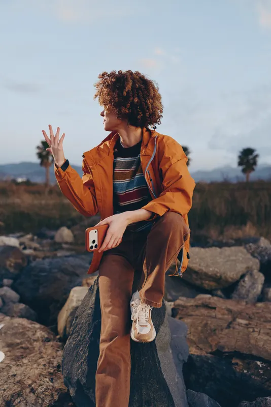 Woman outdoors lifestyle, sitting on rocky stone, curly hair, jacket and backpack, waving hand and holding phone, sunlit mountain landscape, relaxed casual travel journey in daylight