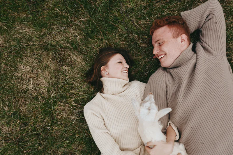 Young couple lying on grass in cozy sweaters smiling at each other while holding a small white baby goat, enjoying a relaxed lifestyle moment outdoors in natural light.