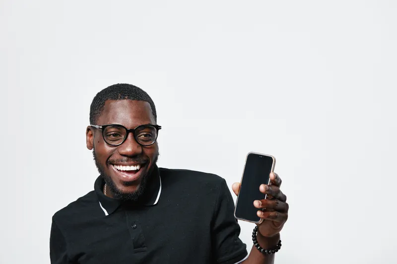 African American man with dark skin and glasses smiling while holding smartphone in hand against white background. Casual black polo shirt and happy expression for technology use.