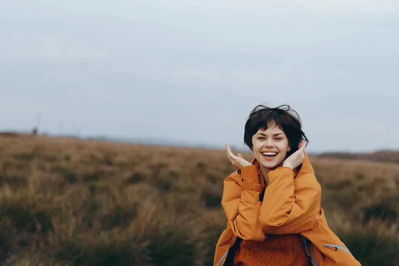 Young woman lifestyle outdoors, smiling with headphones, orange jacket, standing in grassy field under cloudy sky, relaxed joyful moment, soft wind, natural colors, countryside background