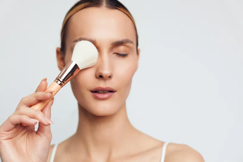 Young woman with light skin applies powder makeup on her closed eye using a large white brush against a plain light background.