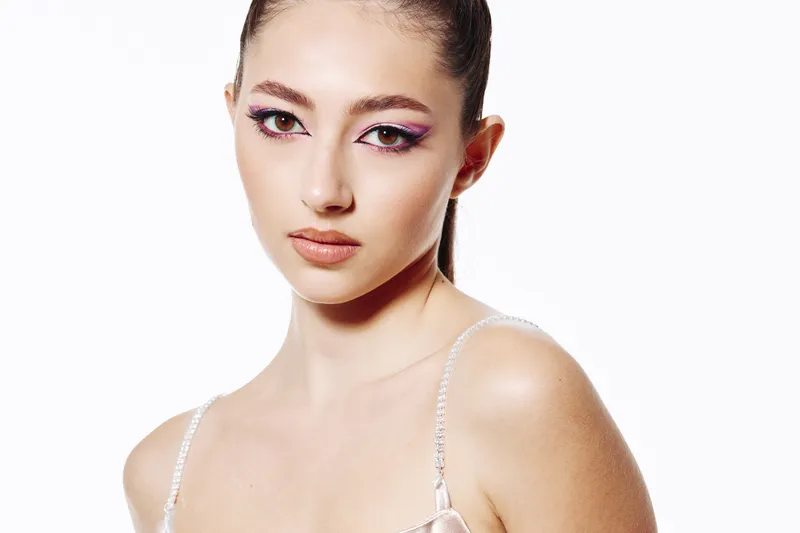 Young woman with dramatic purple glitter eye makeup poses against white. The beauty and fashion portrait emphasizes creative styling and glowing skin. Studio shot high key.