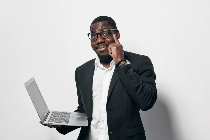 African American man with dark skin wearing glasses and black suit holding laptop and pointing finger to head with confident expression on white background.