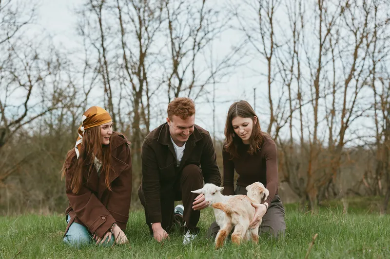 Lifestyle scene with three adults outdoors in a grassy field, interacting with playful goat kids, sharing smiles, and enjoying a candid nature moment together