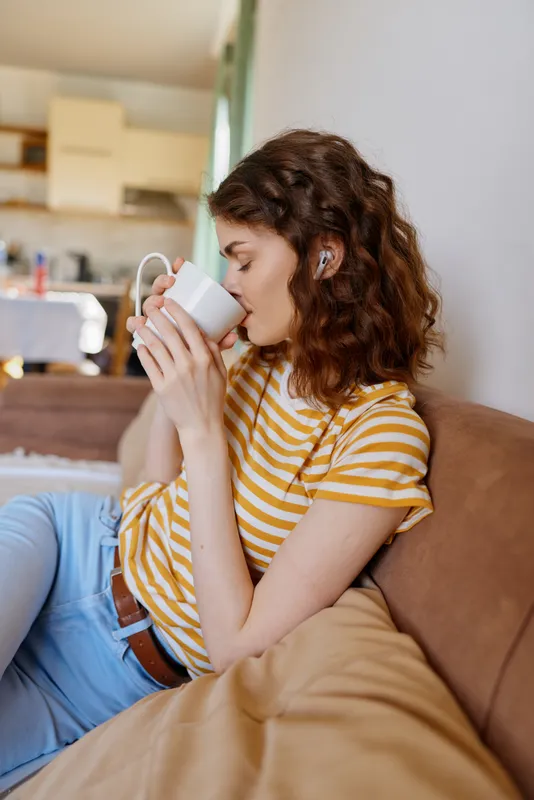 Young woman relaxing at home, sipping from a mug on a sofa, striped yellow and white tee, blue jeans, cozy morning mood.