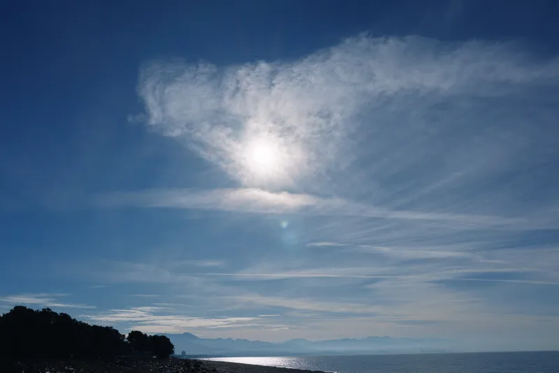 Bright sky over calm sea with a bright sun and wispy clouds, distant coastline silhouette, tranquil coastal landscape under clear weather.