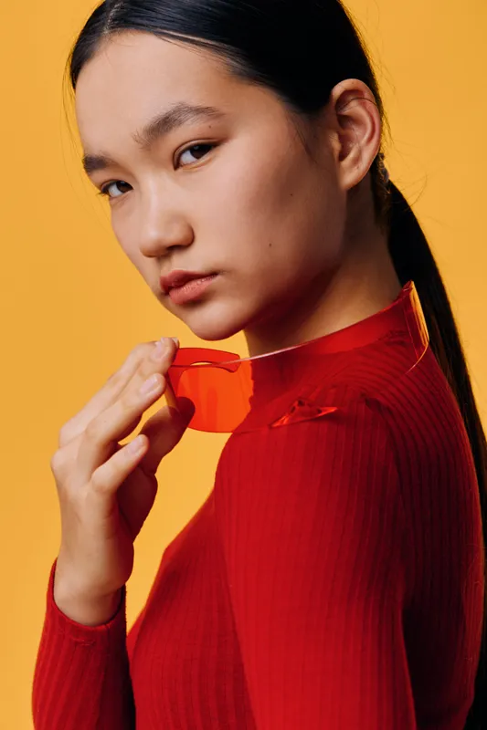 Young woman with black hair wearing red sweater holding red transparent sunglasses near face with serious expression on yellow background in studio portrait fashion style.