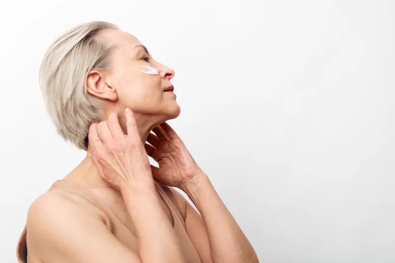 Mature woman with short blonde hair touching her neck and face, eyes closed, against white background. Concept of natural beauty, aging skin care, and self-awareness.