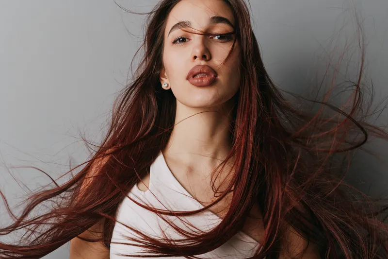 Young woman with long flowing hair and natural makeup posing against gray background. Female model with serious expression in casual white shirt looking at camera.