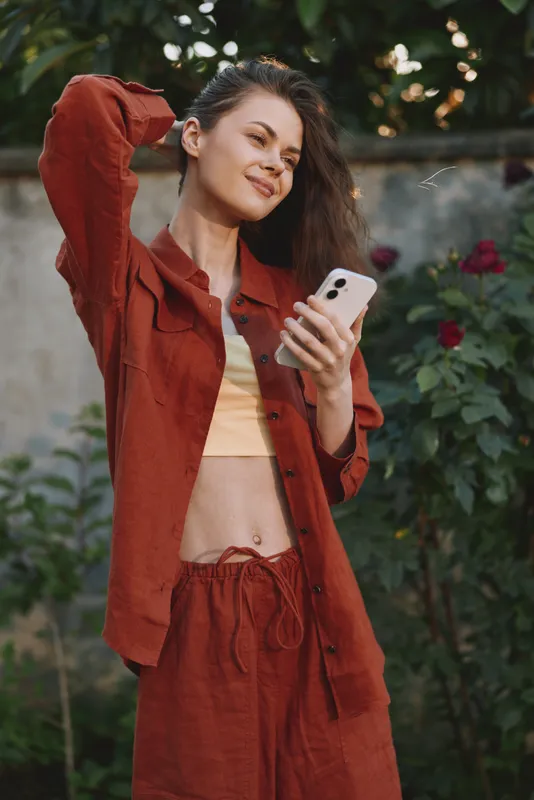 A young woman in a rust colored outfit enjoys a casual moment in a garden as she smiles while talking on her phone, the relaxed pose, soft light and blooming roses creating a warm lifestyle vibe