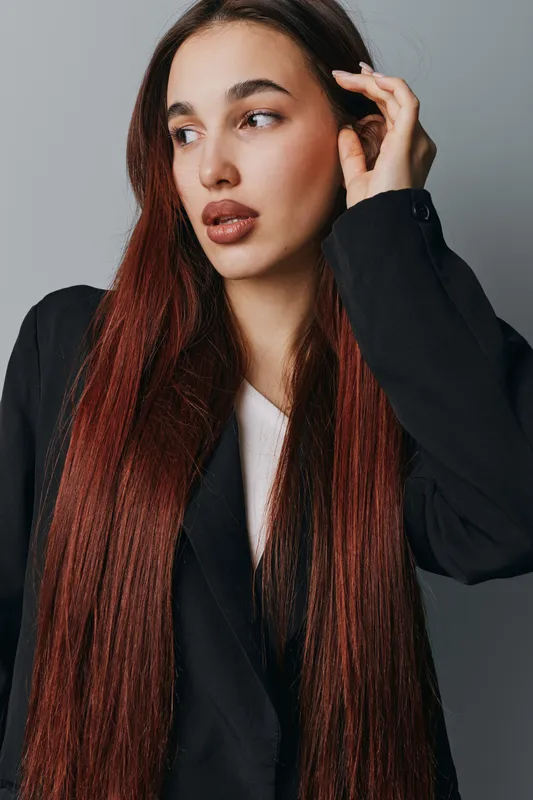 Woman with long red hair wearing black blazer and white shirt posing with hand near face against gray background. Portrait of confident female model with natural makeup and serious expression.