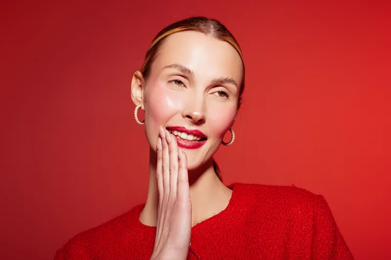 Smiling woman with red lipstick and earrings touches her face on red background. Portrait of happy model with natural skin and shiny makeup. Concept of beauty, fashion, and positive emotions.