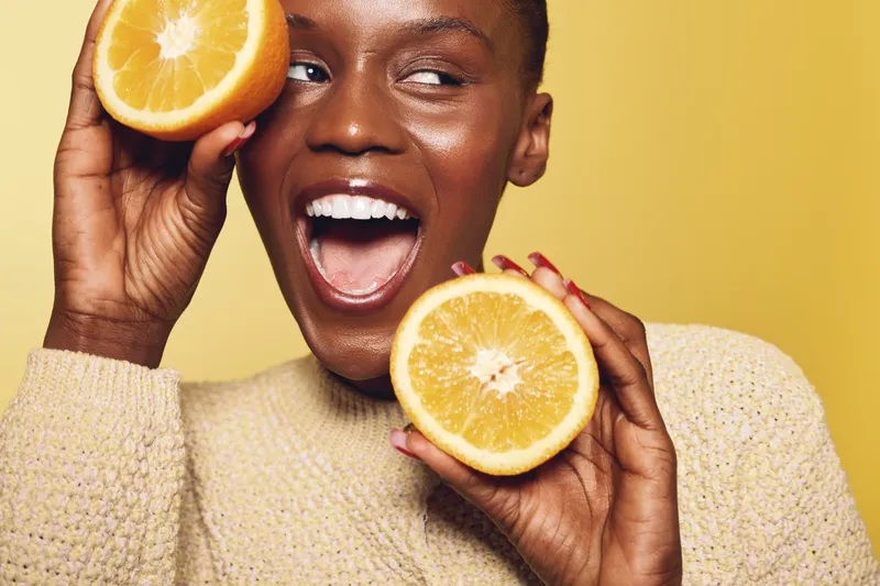 Happy African woman with dark skin holding two orange halves near her face smiling with open mouth on yellow background. Concept of healthy lifestyle, fruit, and natural beauty.