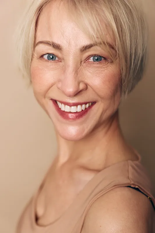 Close-up portrait of a smiling middle aged woman with short blonde hair and blue eyes wearing a beige top. Natural beauty concept with positive expression and healthy skin in soft lighting.