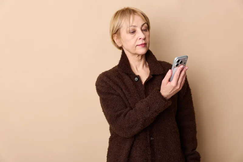 Elderly woman smartphone texting messaging communication technology portrait indoors beige, senior female in warm coat, holding phone typing, calm focused
