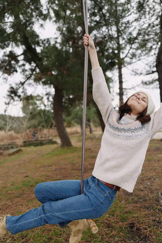 Girl on a swing in a park, enjoying outdoor play, wearing a cozy sweater and jeans, arms raised, smiling as she soars among trees and earthy ground.