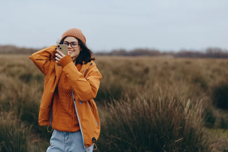 Smiling woman in a field outdoors, with a smile, wearing a knit hat and orange jacket, holding a warm drink near her mouth, autumn lifestyle in tall grass, natural light, calm mood