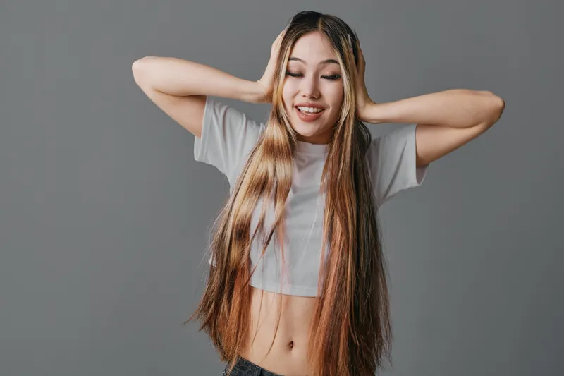 Young woman with long hair covering ears, smiling with eyes closed on gray background. Casual clothing, relaxed pose and fun expression, female person with natural beauty and happiness.