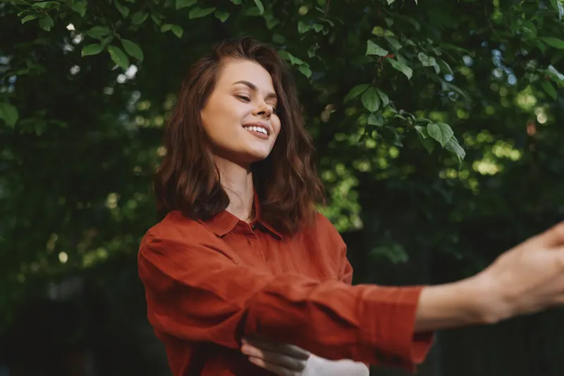 Natural woman lifestyle portrait outdoors, smiling woman in rust shirt, relaxed pose among green leaves, candid moment of joy, sunny day, casual creative scene, upbeat mood