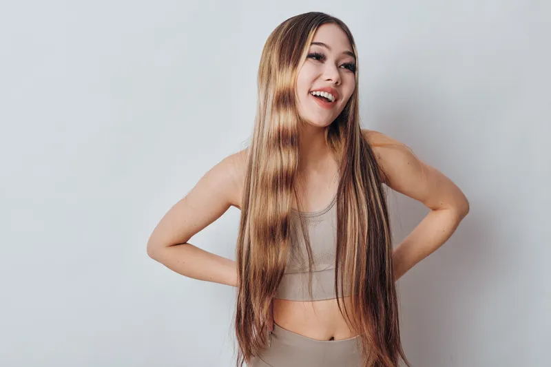 Young woman with long straight hair in casual top smiling and posing on light background. Female model looking away happy and confident with natural makeup and relaxed posture.
